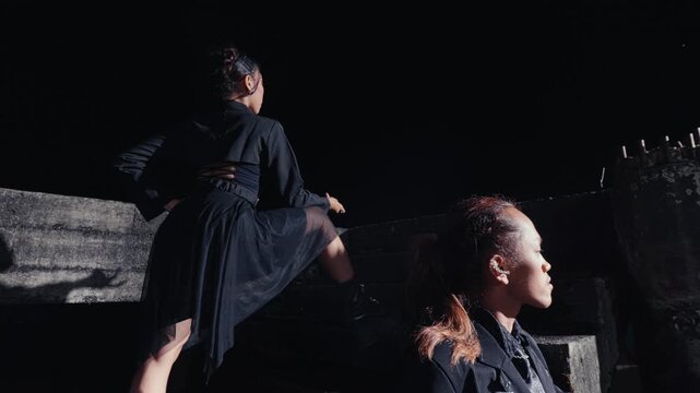 A fashion-forward man and woman in all-black outfits pose on concrete stairs in a dark, industrial setting at night. The camera pulls back, revealing their stylish and confident presence.