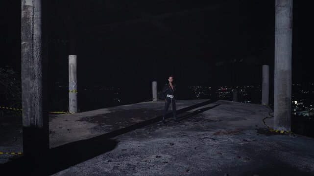 A stylish model in an all-black outfit stands alone in a derelict building, posing with confidence against a vast backdrop of sparkling city lights at night. A shot of urban solitude and power.