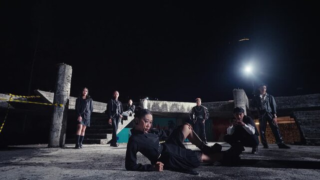 A group of models in edgy black outfits poses dramatically in an abandoned concrete building. A person in the foreground enters the frame, adding a dynamic layer to this stylish, urban night scene.