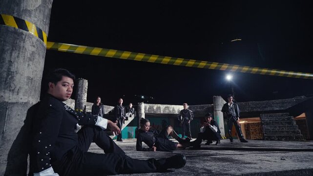 A stylish man in a designer jacket sits against a concrete pillar, with his edgy crew posing behind him in an abandoned building. A cool, confident shot of a leader and his gang.