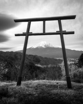 View of Mount Fuji framed by a traditional wooden Torii gate overlooking forested hills and a valley under a cloudy sky Fujinomiya, Shizuoka, Japan.