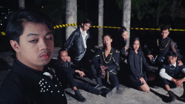 A stylish young man poses for a selfie video, looking seriously at the camera, while his fashion-forward gang sits and poses in the background of a derelict, industrial location at night.