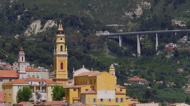 Aerial sweep over Menton, France, shows the Basilica of Saint Michael, tall bell tower, pastel houses, palm promenade, docks, and a concrete viaduct at midday.