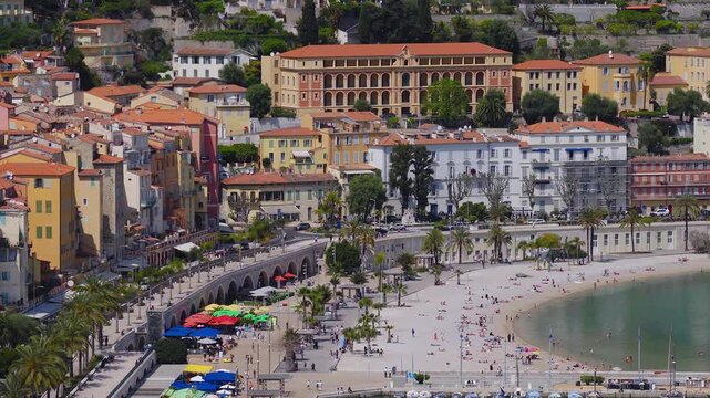 Aerial view of Menton on the French Riviera shows the old town, arched viaduct, palm promenade, market umbrellas, and beachgoers under bright midday sun.