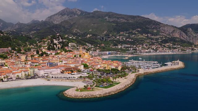 Aerial view of Menton, French Riviera, shows the crescent old town, yellow bell tower, beach, breakwater marina, and Maritime Alps in bright midday light.