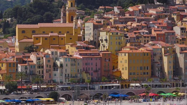 Aerial view of Menton on the French Riviera shows the Basilica of Saint Michel, terracotta roofs, arcaded quay, marina masts, palm lined promenade, and beach umbrellas.