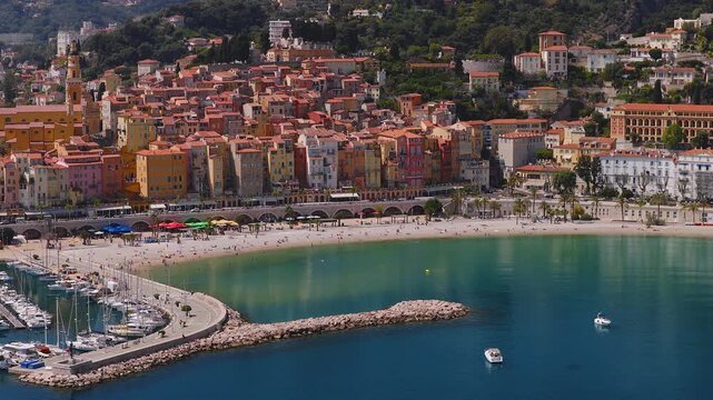 Aerial view of Menton on the French Riviera shows a crescent bay, sandy beach, marina, church bell tower, arched viaduct, curved breakwaters, boats, and promenade.