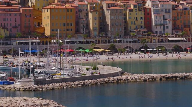 Aerial view of Menton, France shows the old quarter behind a curved marina and sandy shore, with arched stone promenade, palm seafront, sailboats, and people strolling and dining.