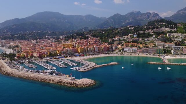 Aerial view of Menton, France shows a curved marina, long rock breakwater, moored yachts, old town with yellow church tower, palm promenade, and crescent beach.