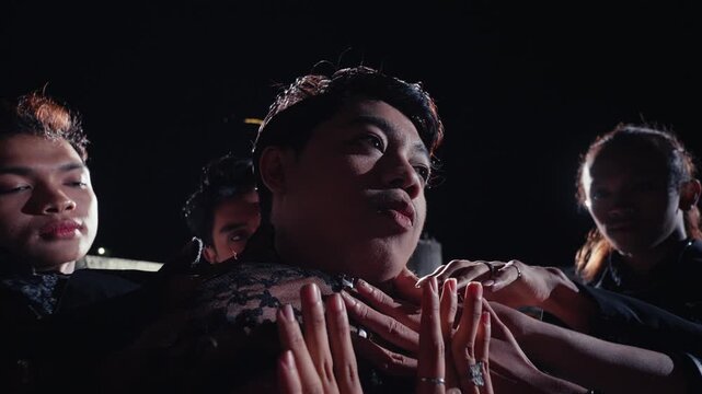 A close-up of a young man's face being held by multiple hands from his group. The camera pulls back to reveal the full, intense scene of a cult-like ritual on a dark rooftop.