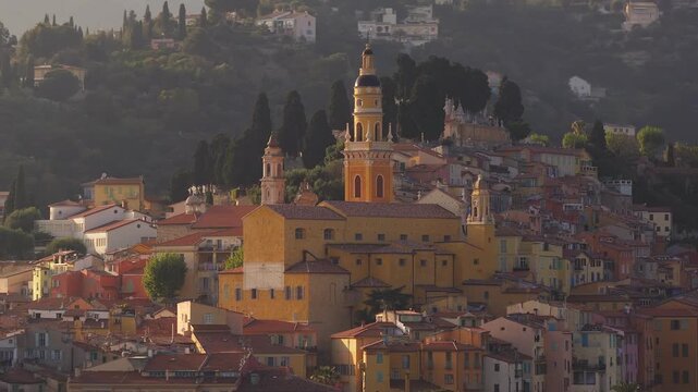 Aerial view of Menton, French Riviera, with pastel facades and terracotta roofs. Camera pans across hillside, cypress rows, and the ochre bell tower at golden hour.