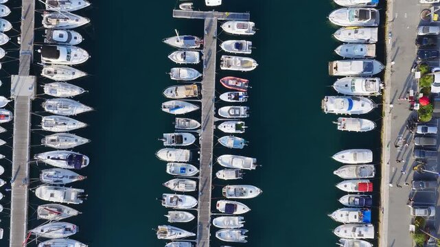 Aerial view of Menton, France marina with parallel piers, moored boats, quay walkers, umbrellas, parking, late afternoon sun, and submerged quay structures visible.