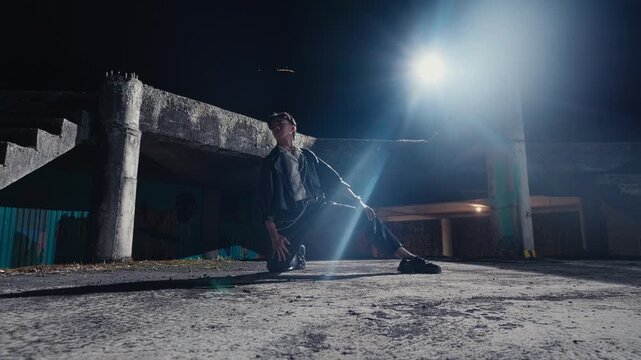 A young man in a leather jacket and stylish clothes strikes a powerful pose alone in a derelict concrete building at night, dramatically backlit by a bright, flaring light.