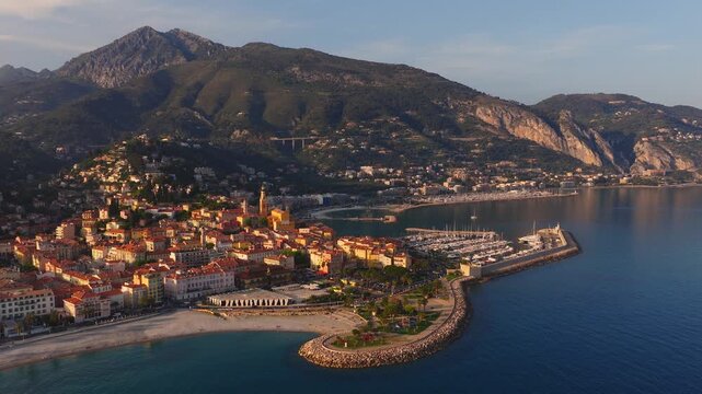 Aerial glide over Menton on the French Riviera shows old town ochre blocks, church bell tower, marina, curved breakwater, yachts, beachgoers, palms, and rocky hills.