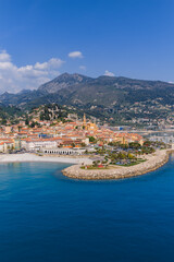 Fototapeta premium Aerial view of Menton, France shows pastel Old Town, Basilique Saint Michel bell tower, curved stone jetty, marina, Promenade du Soleil, turquoise sea, and Maritime Alps at midday.