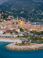 Fototapeta premium Aerial Menton, France, shows the Basilica of Saint Michel bell tower, terracotta roofs, marina and promenade, curved breakwater, sandy beach, and Alpine foothills in sun.