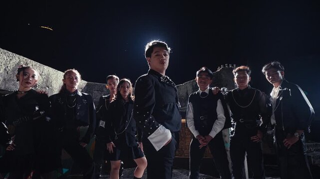 A diverse group of young models in edgy black outfits poses together with confidence on a rooftop at night, strongly backlit by a spotlight for a dramatic, cinematic effect.