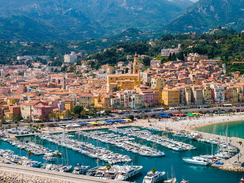 Aerial view of Menton, France, shows pastel old town, Basilique Saint Michel tower, marina of white yachts and sailboats, turquoise water, promenade, and Maritime Alps.
