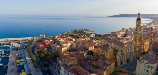 Naklejka premium Aerial view of Menton, France, with Basilica of Saint Michel Archange, Promenade du Soleil, pebbled beach, marina, market tents, and Cap Martin in early evening light.