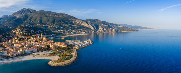Obraz premium Aerial view of Menton, France, shows Basilica of Saint Michel tower, pastel old town, marina, breakwater, Maritime Alps and bays in clear morning light and calm water.