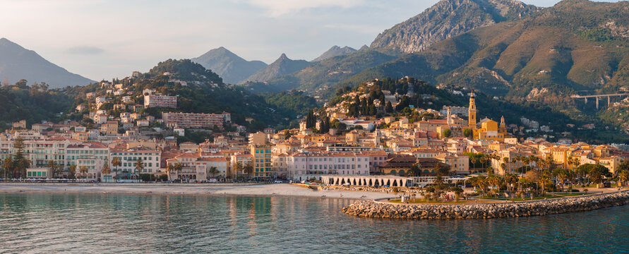 Aerial view of Menton, France, with the Basilica of Saint Michel, crescent promenade, curved breakwater, calm harbor, and hillside old town in late afternoon light.