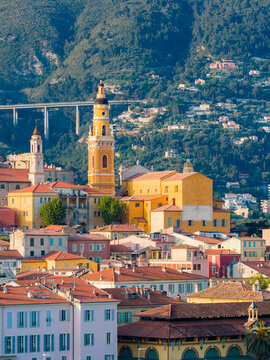 Menton, France hillside shows the Basilica of Saint Michel bell tower, stacked pastel old town, tiled roofs, a sweeping viaduct, rugged mountains, and warm daylight.