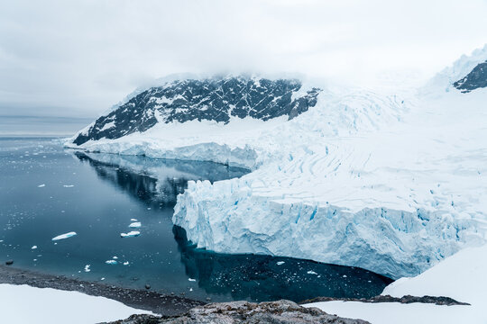 View of Neko Harbor featuring a massive glacier wall meeting dark blue water with floating ice and snow-capped mountains under a cloudy sky in Antarctica.