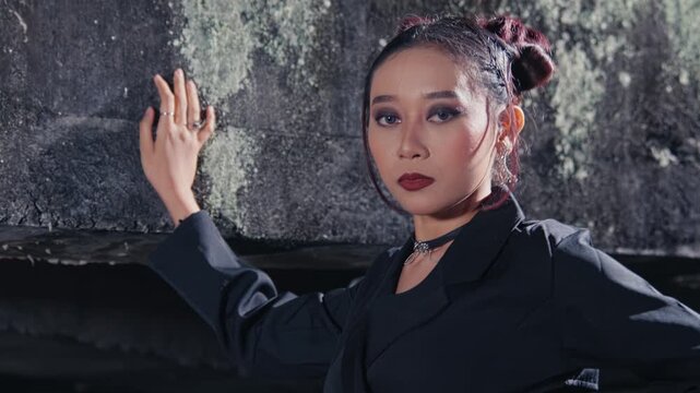 A stylish woman with dramatic makeup and a choker necklace poses with confidence against a rough stone wall at night, looking directly at the camera with a fierce and alluring gaze.