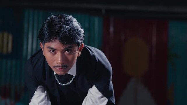 A young man with a mustache and black painted nails leans forward, staring intensely at the camera in a dark, urban location. A powerful and edgy portrait with a rebellious attitude.