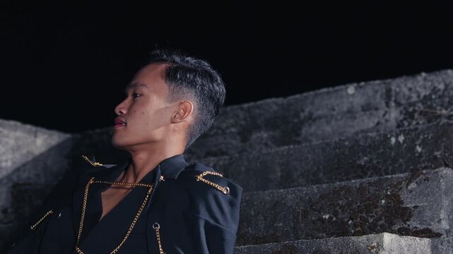 A handsome young man in a stylish black jacket with gold chains sits on concrete stairs at night, posing for a fashion shoot with a cool and confident expression.