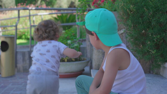 Toddler stumbling and falling while playing near older boy sitting on stone steps in garden with flowers and greenery in the background