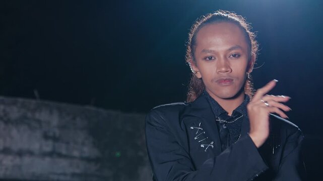 A young man with long hair, earrings, and a punk-inspired jacket with safety pins poses for the camera, backlit by a strong light that creates a dramatic lens flare.