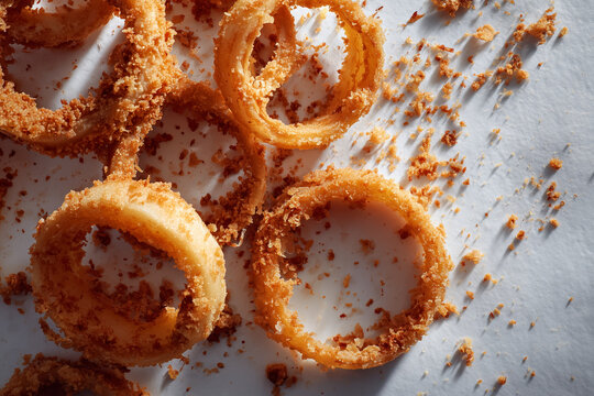 Crispy onion rings arranged with messy crumbs around on light background.