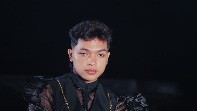An androgynous model with a stylish haircut wears a sheer black shirt and vest, posing with a serious and confident expression against a dark background at night.