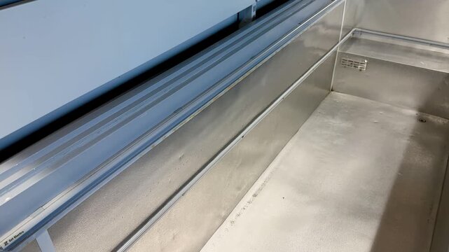 Empty industrial freezer interior stainless trough cleaned after defrost, metal rails and drainage channel visible, cool blue