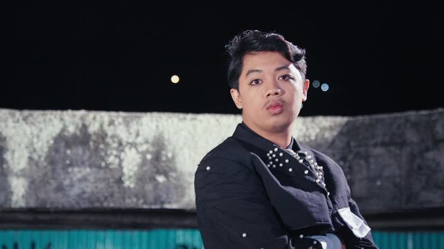 A young man in a stylish jacket with pearl studs poses in a gritty, urban location at night, with a graffiti-covered wall behind him. He looks at the camera with a cool, confident expression.