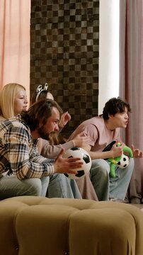 Friends joyfully react while watching a soccer game on a couch, holding soccer balls and showing excitement in a stylish living room
