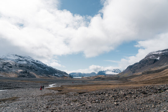 View of the rugged valley and snow-capped mountains with people walking across the rocky terrain under a cloudy sky in Stromness, South Georgia and the South Sandwich Islands.