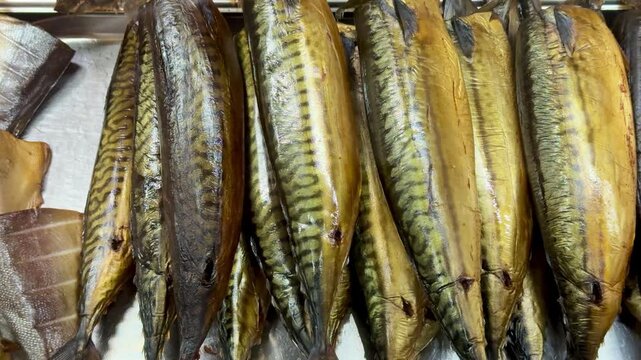 Rows of cold smoked mackerel on seller counter, golden oily skins glisten under warm lighting, fishmonger arranges fillets