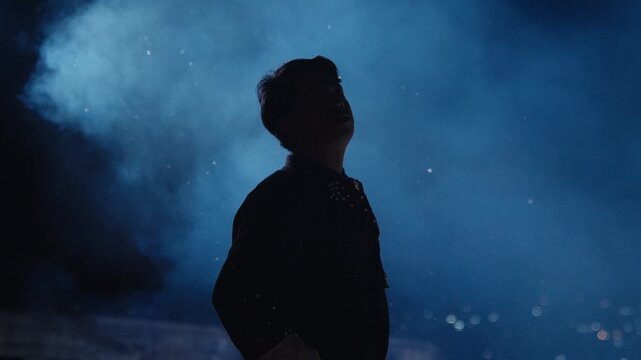 A man's silhouette is captured in a moody, atmospheric setting with blue smoke and a strong backlight. He slowly turns his head, looking out into the distance, conveying a sense of thought or wonder.