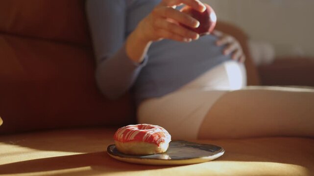 Expectant mother making a healthy food choice, picking a fresh red apple instead of a sugary donut for a snack while sitting on the couch and lovingly caressing her pregnant belly