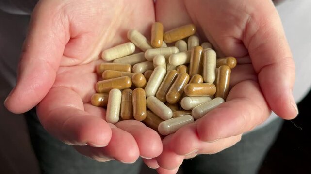Women's hands filled with brown and beige capsules containing various medications, close-up capsules illuminated by warm sunlight