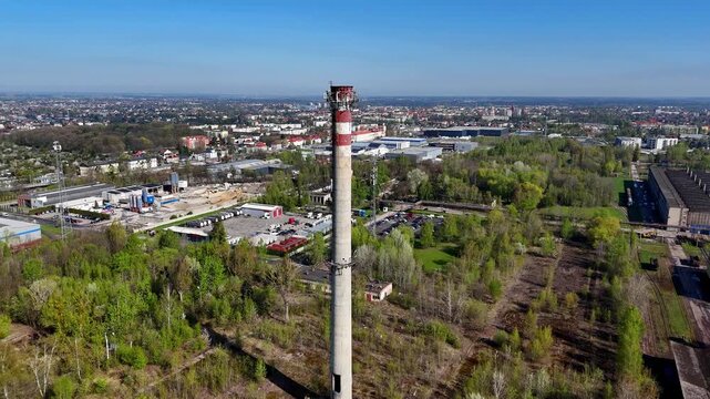 Aerial drone orbital shot of a tall concrete industrial smokestack with a red and white top section, equipped with telecommunication antennas. Background features urban cityscape, trees, warehouses, p