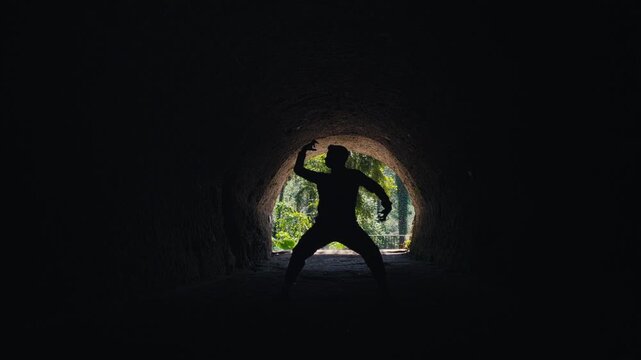 A slow dolly push-in shot of a man's powerful silhouette at the end of a dark cave. The movement enhances the feeling of approaching a goal, freedom, or a new beginning.