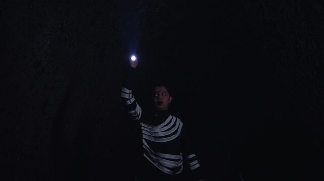 A man with a terrified expression and dramatic makeup explores a pitch-black cave, waving a flashlight around. The scene is perfect for horror, suspense, adventure, or mystery themes.