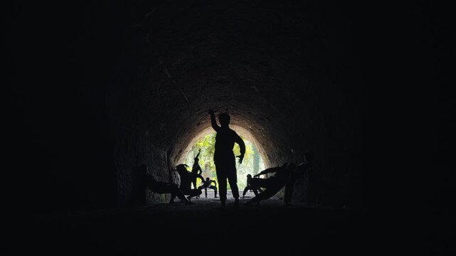 A wide, static shot showing the silhouettes of a group of dancers or performers. One person stands as a leader while others are posed around them at the light-filled end of a dark cave.