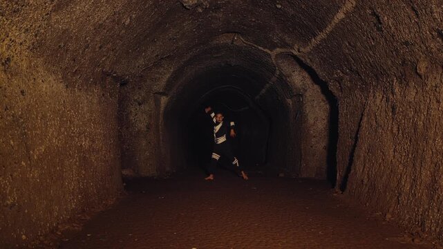 A barefoot man in a unique costume performs an intense, ritualistic martial arts dance with a rope or whip inside a dark, ancient cave. The scene is mystical, powerful, and culturally rich.