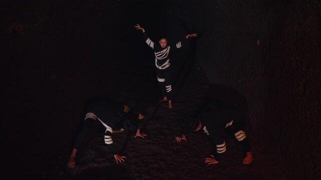 A high-angle shot captures three performers in a dark cave. A woman dances expressively while two men stay in low, primal poses, creating a mysterious and ritualistic atmosphere.