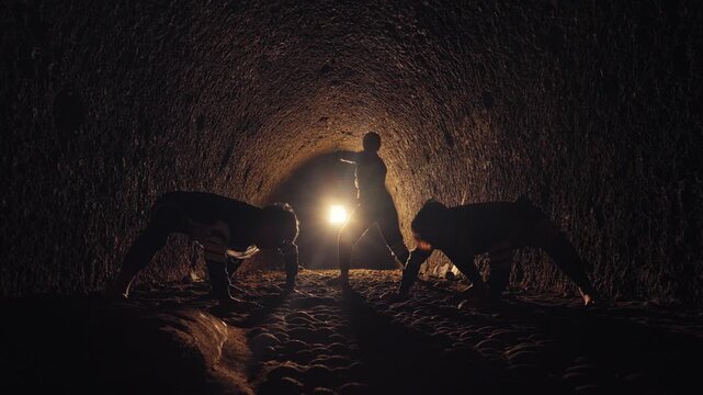 A powerful low-angle shot of three dancers silhouetted in a dark tunnel. A woman stands and moves mystically while two men crouch, backlit by a single bright light, creating a spiritual scene.
