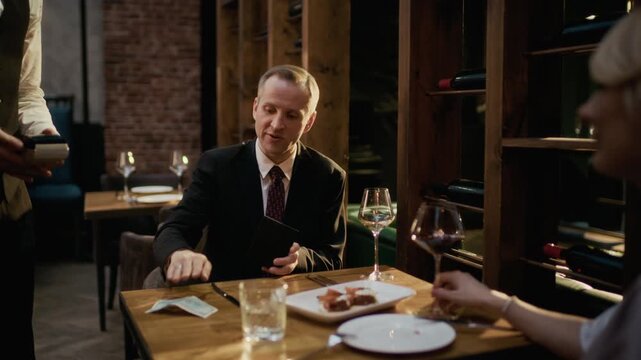 Handheld medium shot of middle-aged Caucasian male guest wearing suit calling waiter and paying for restaurant dinner in cash, then clinking glasses with blonde woman in foreground